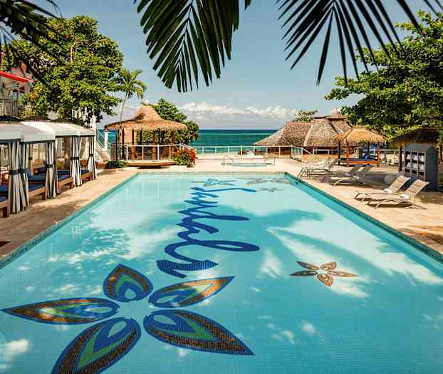 En pool med omgivande solstolar och en byggnad med halmtak synlig vid havet. (AI-genererad text)