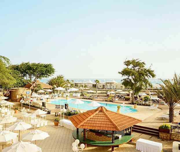 Bild av ett utomhusområde vid en pool med parasoller och träd under klar himmel. (AI-genererad text)