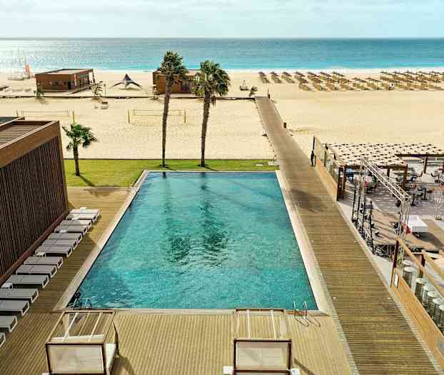 Utomhuspool med solstolar, gräsmatta, strand med parasoller och havet i bakgrunden. (AI-genererad text)