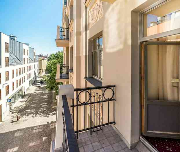 Bilden visar en balkong på en ljus byggnad med utsikt över en stenlagd gata och ett modernt hus med stora fönster på andra sidan gatan. (AI-genererad text)