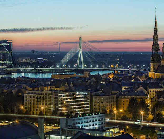 Panoramabild av en stad i skymningen, med en flod, upplysta byggnader och en bro i bakgrunden. (AI-genererad text)