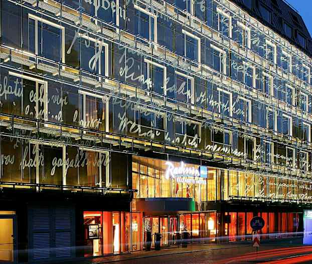 En modern byggnad med glasfasad upplyst på kvällen, dekorerad med skrivstilar. Ingången till en hotellkedja syns tydligt. (AI-genererad text)