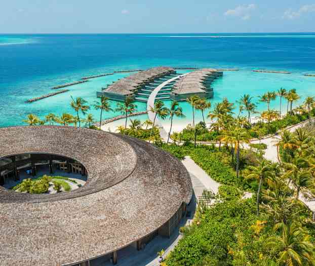 Vy över en rund byggnad med tak av trä och vattenvillor omgivna av turkost hav. (AI-genererad text)