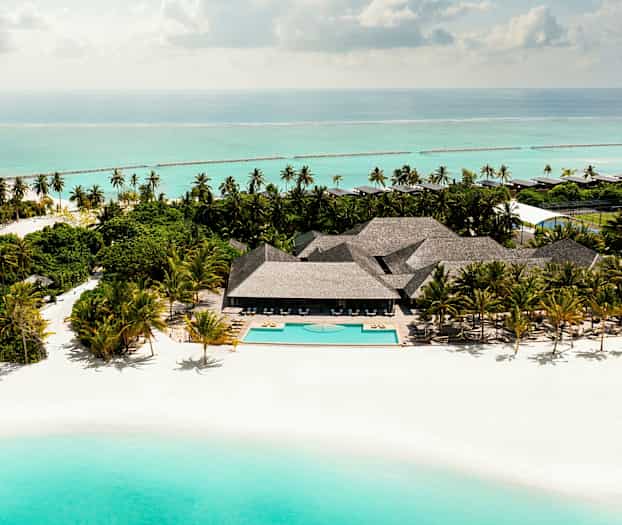 Flygbild av en sandstrand med en pool, omgiven av palmer och byggnader vid havet. (AI-genererad text)