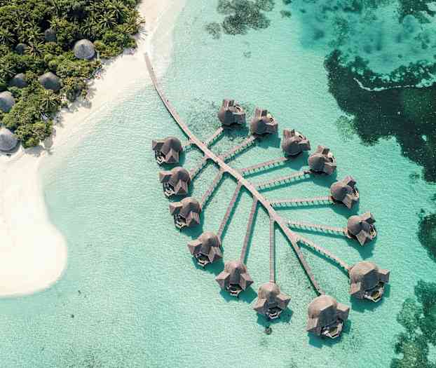 Flygbild över bungalows på pålar över klarblått vatten med en närliggande sandstrand och frodig vegetation. (AI-genererad text)