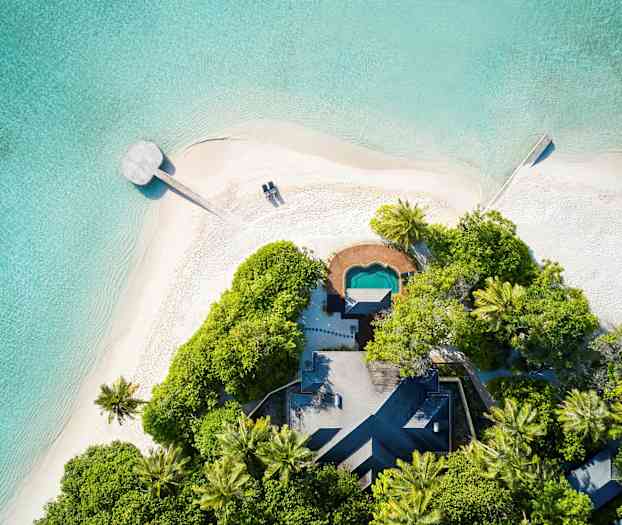Flygbild av sandstrand vid kristallklart tropiskt hav, med grönskande vegetation och en liten byggnad. (AI-genererad text)