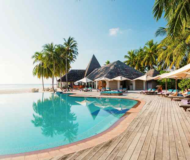 En infinitypool med träd och parasoller vid sidan, samt byggnader med halmtak i bakgrunden. (AI-genererad text)