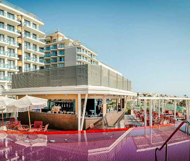 Ett hotell med balkonger och en pool framför, samt en bar och sittplatser under parasoller. (AI-genererad text)