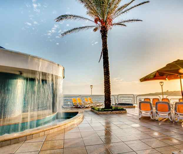 En pool med ett vattenfall intill en palm, solstolar och utsikt över havet under en solig dag. (AI-genererad text)