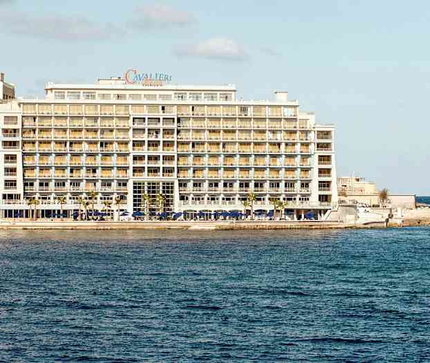 Ett stort hotell vid havet, med flera våningar och balkonger samt en liten båt i vattnet nära hotellet. (AI-genererad text)