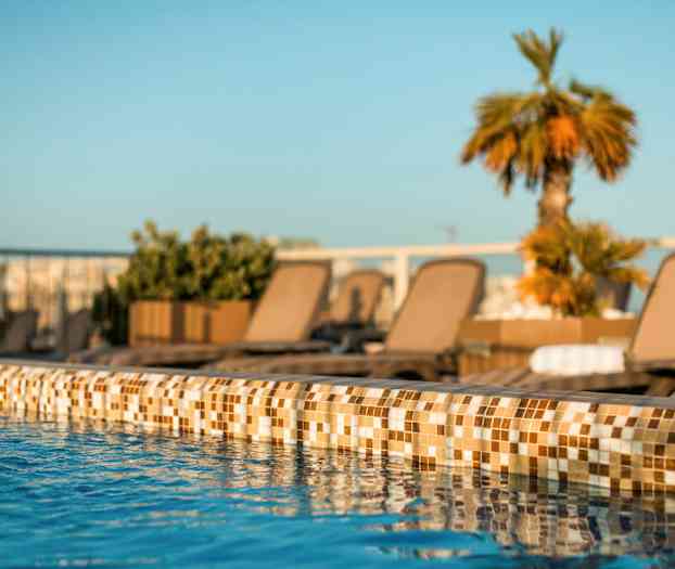 Närbild av en poolkant med klarblått vatten och solstolar samt växter i bakgrunden. (AI-genererad text)