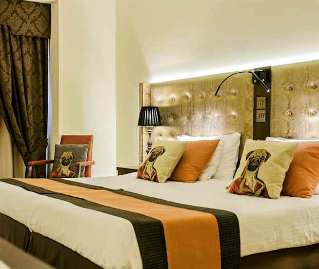 Ett hotellrum med en stor bäddad säng, dekorerad med kuddar med hundmotiv. Rummet har mörka gardiner och en stol med matchande kudde. (AI-genererad text)