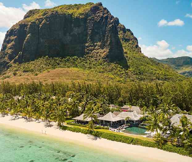 Ett stort berg syns i bakgrunden av en strand med turkost vatten i förgrunden. På stranden står palmträd och byggnader med halmtak. (AI-genererad text)