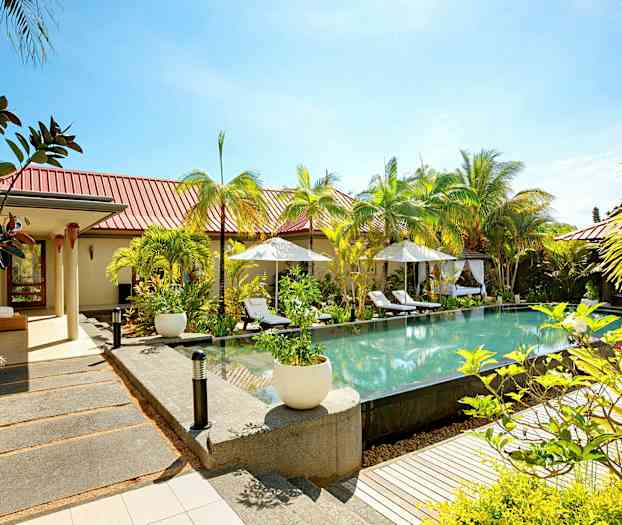 En öppen miljö med en pool omgiven av solstolar och parasoller, med grönska och byggnader i bakgrunden. (AI-genererad text)