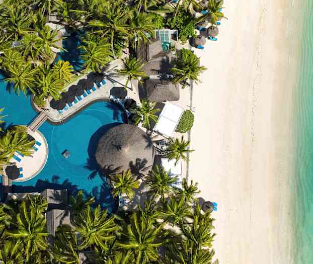 Bilden visar en flygvy över en strand med vit sand, turkost vatten, och en pool omgiven av palmer. (AI-genererad text)