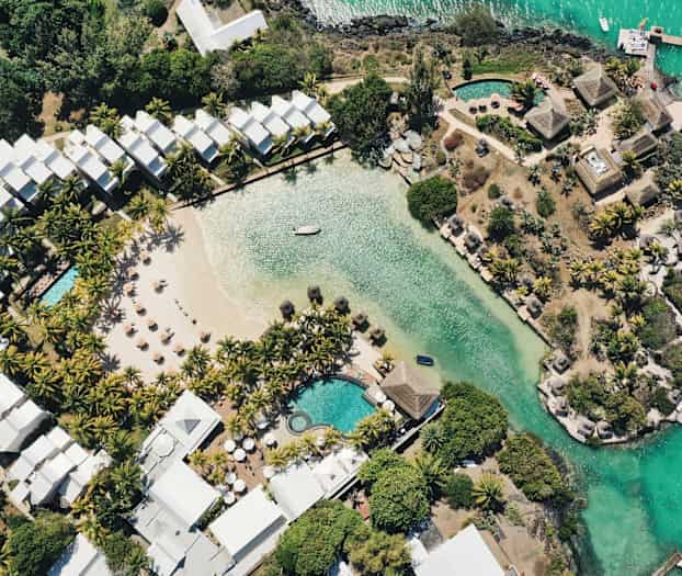 Flygfoto av ett hotellområde med pool, en sandstrand och grönska vid vattnet. (AI-genererad text)