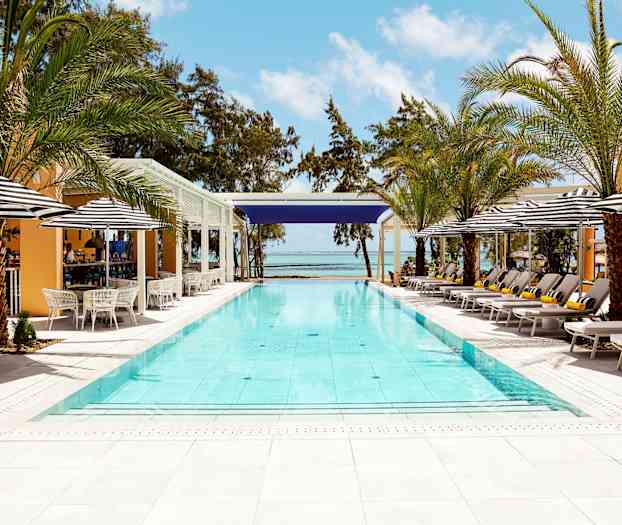 Utomhuspool omgiven av solstolar, parasoller och palmer med hav och blå himmel i bakgrunden. (AI-genererad text)