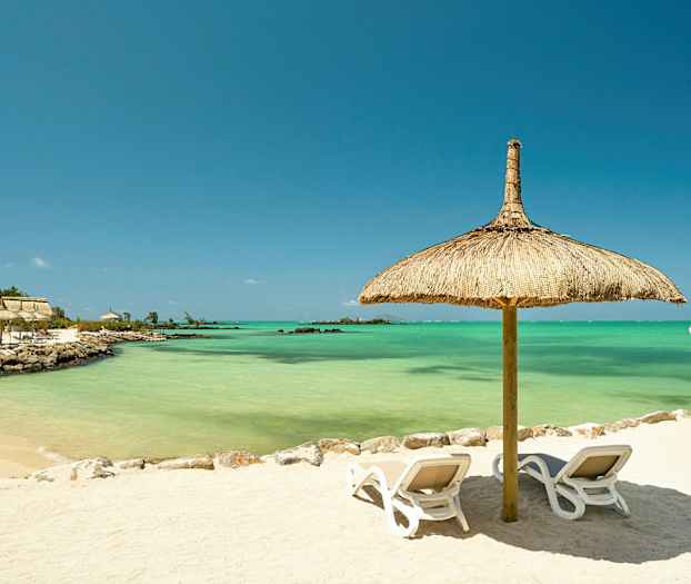 En sandstrand med två solstolar under ett parasoll av strå, med turkost vatten i bakgrunden. (AI-genererad text)