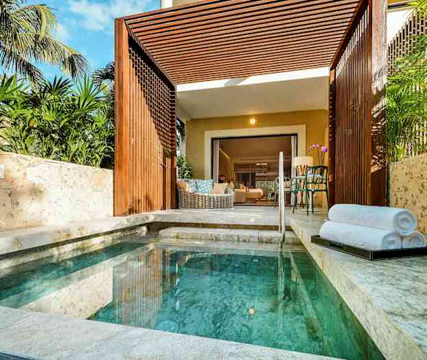 En liten pool omgiven av stenbeläggning och grönska med en öppen uteplats som leder in till ett rum. (AI-genererad text)