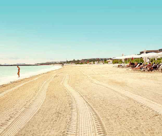 En sandstrand med parasoller och solstolar, en person står vid vattnet. (AI-genererad text)