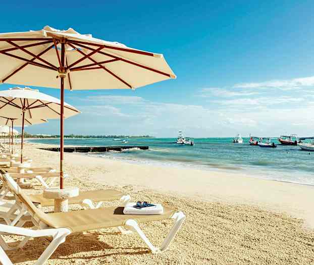 En sandstrand med solsängar och parasoll. I bakgrunden syns havet med båtar. (AI-genererad text)