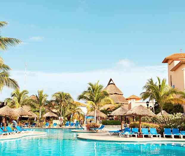 Bilden visar en utomhuspool omgiven av solstolar med blå dynor och parasoller. I bakgrunden syns palmer och byggnader med orangeröda tak. (AI-genererad text)