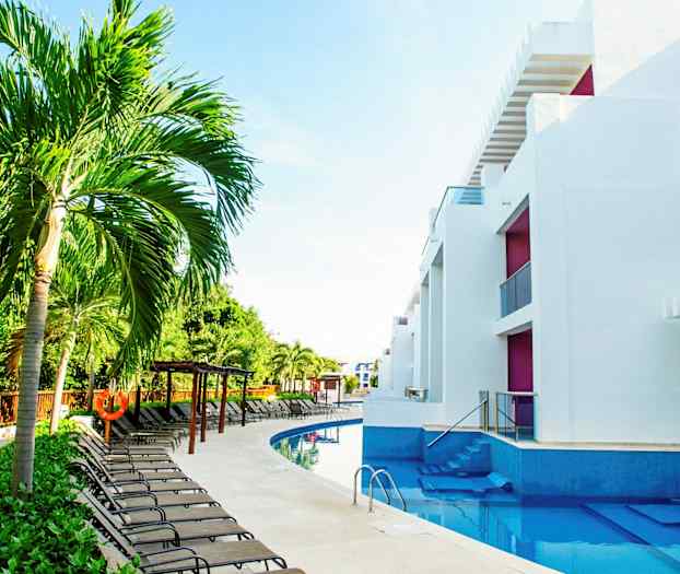 Bild av en swimmingpool omgiven av solstolar, palmer och moderna vita byggnader. (AI-genererad text)