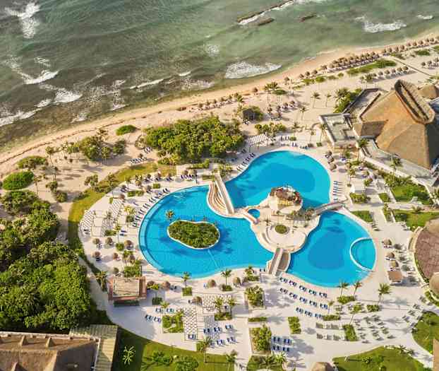 Flygfoto av en semesterort vid havet med flera pooler, omgivande grönska och byggnader med halmtak. (AI-genererad text)