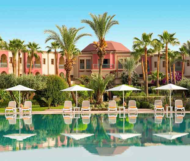 En swimmingpool framför en byggnad omgiven av palmer och parasoller. (AI-genererad text)