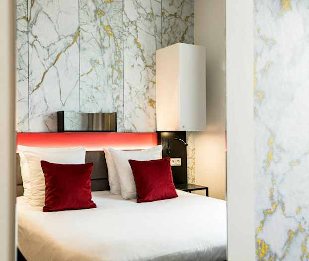 Ett modernt hotellrum med en dubbelsäng, vita och röda kuddar samt en bakgrundsvägg med marmormönster. (AI-genererad text)