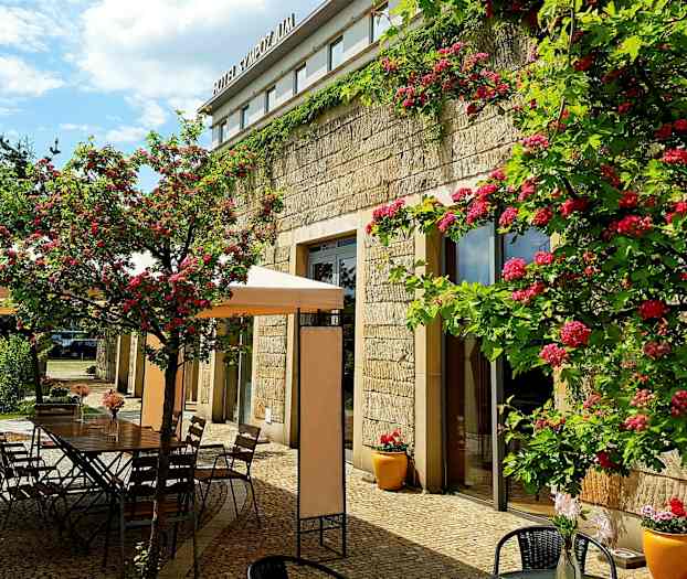 En utomhusterrass med blommor och bord intill en stenbyggnad. (AI-genererad text)