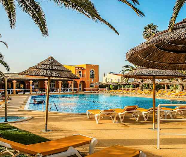 Utomhuspool omgiven av solstolar och halmtakparaplyer, med en byggnad i bakgrunden. (AI-genererad text)