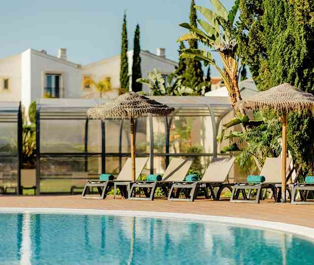 Solstolar och parasoller vid en pool med byggnader och växtlighet i bakgrunden. (AI-genererad text)