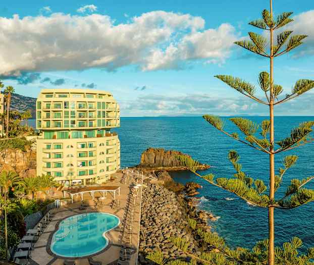 Ett hotell vid kusten med en pool och omgivet av hav och träd. (AI-genererad text)