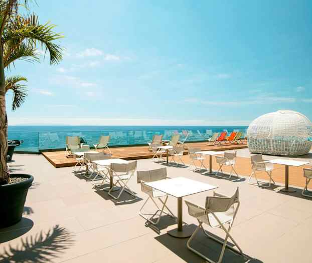 En utomhusterrass med vita stolar och bord, skuggande palmer, utsikt över havet samt en vit kupolformad struktur i bakgrunden. (AI-genererad text)