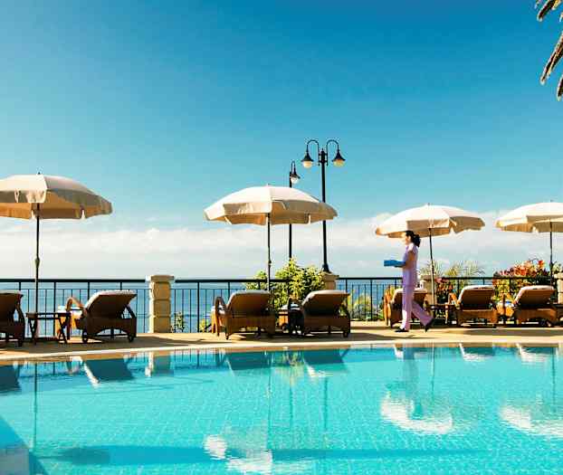 En utomhuspool med solstolar och parasoller vid sidan, samt en person som bär en bricka. (AI-genererad text)