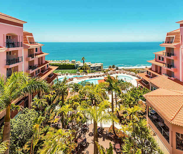 Vy över hotellområde med rosa byggnader, palmer och en pool med solstolar nära havet. (AI-genererad text)