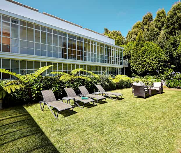 Bild av en trädgård med solstolar, sittgrupp och gröna växter framför en byggnad med stora fönster. (AI-genererad text)