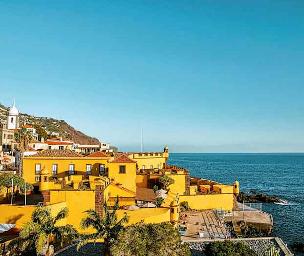 Gul byggnad vid havet med omgivande grönska och kullar i bakgrunden. (AI-genererad text)