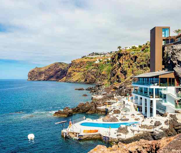 Kustlandskap med en modern byggnad och poolområde vid klipporna, omgiven av havet och grönskande sluttningar. (AI-genererad text)