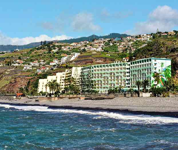 Hotellbyggnader vid en strand med havsvågor och en bakgrund av gröna kullar och bostadshus. (AI-genererad text)