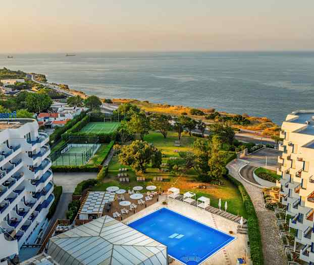 Utsikt över ett hotellområde vid havet med pool, grönområden, och sportplaner. (AI-genererad text)