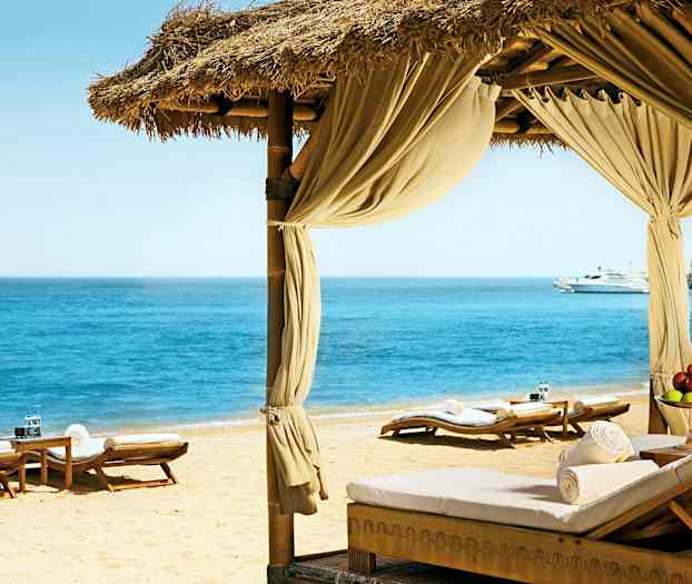 En strand med solstolar och en paviljong ser ut mot ett klart hav. (AI-genererad text)