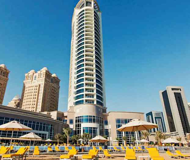 Bilden visar en modern hög byggnad omgiven av flera andra byggnader och ett område med gula solstolar och parasoller. (AI-genererad text)