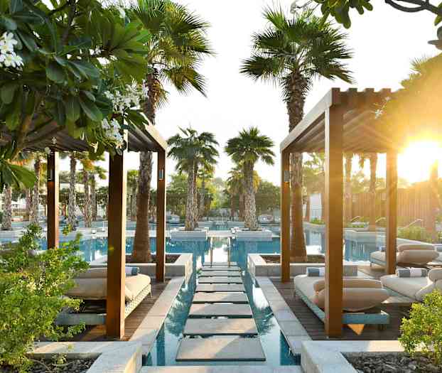 En trädgårdsanläggning med pooler, solstolar och palmer under ett tak av pergolor med solsken i bakgrunden. (AI-genererad text)