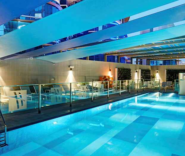 En pool inomhus med belysning. Runt poolen finns vilstolar och glasräcken. (AI-genererad text)