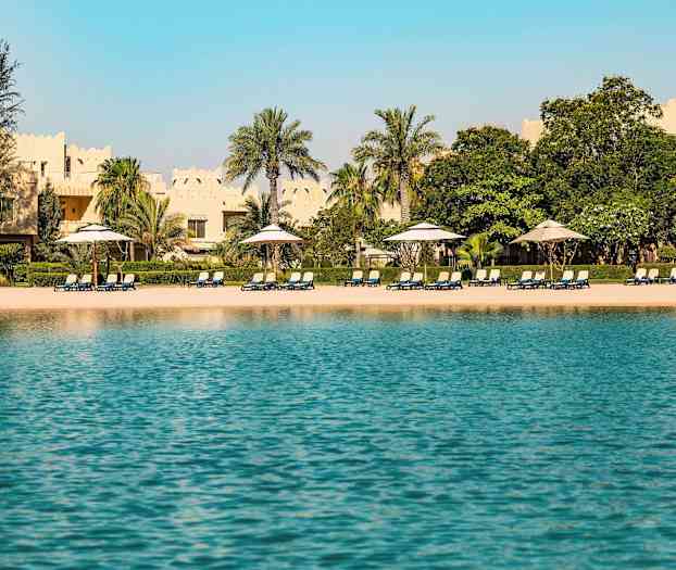 Utsikt över en strand med solstolar och parasoller, omgiven av palmer och byggnader med traditionell arkitektur i bakgrunden. (AI-genererad text)