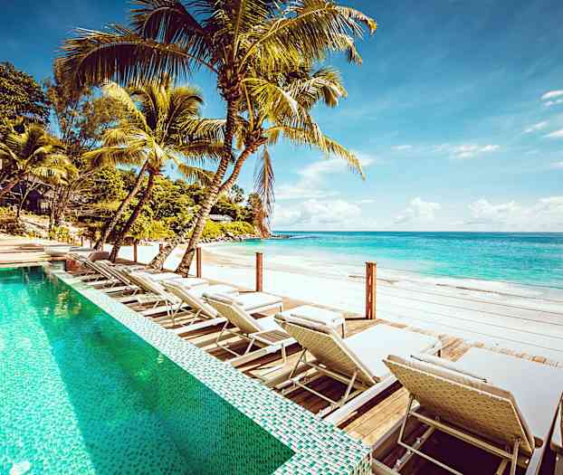 Utomhuspool med solstolar vid en sandstrand med klart blått vatten. (AI-genererad text)