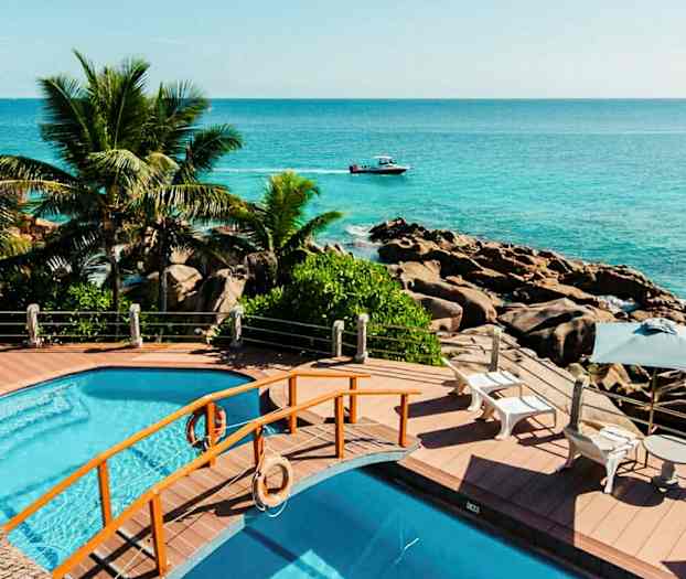 Poolområde med utsikt över havet, omringad av palmer och klippor. (AI-genererad text)