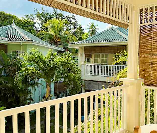 En vy från en balkong som visar ett grönskande område med pastellfärgade hus och palmträd. (AI-genererad text)